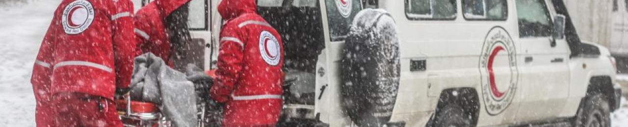 Four Syrian Red Crescent staff, wearing winter jackets with the hood pulled up, lift a stretcher into the back of a Red Crescent landrover during a snowstorm.